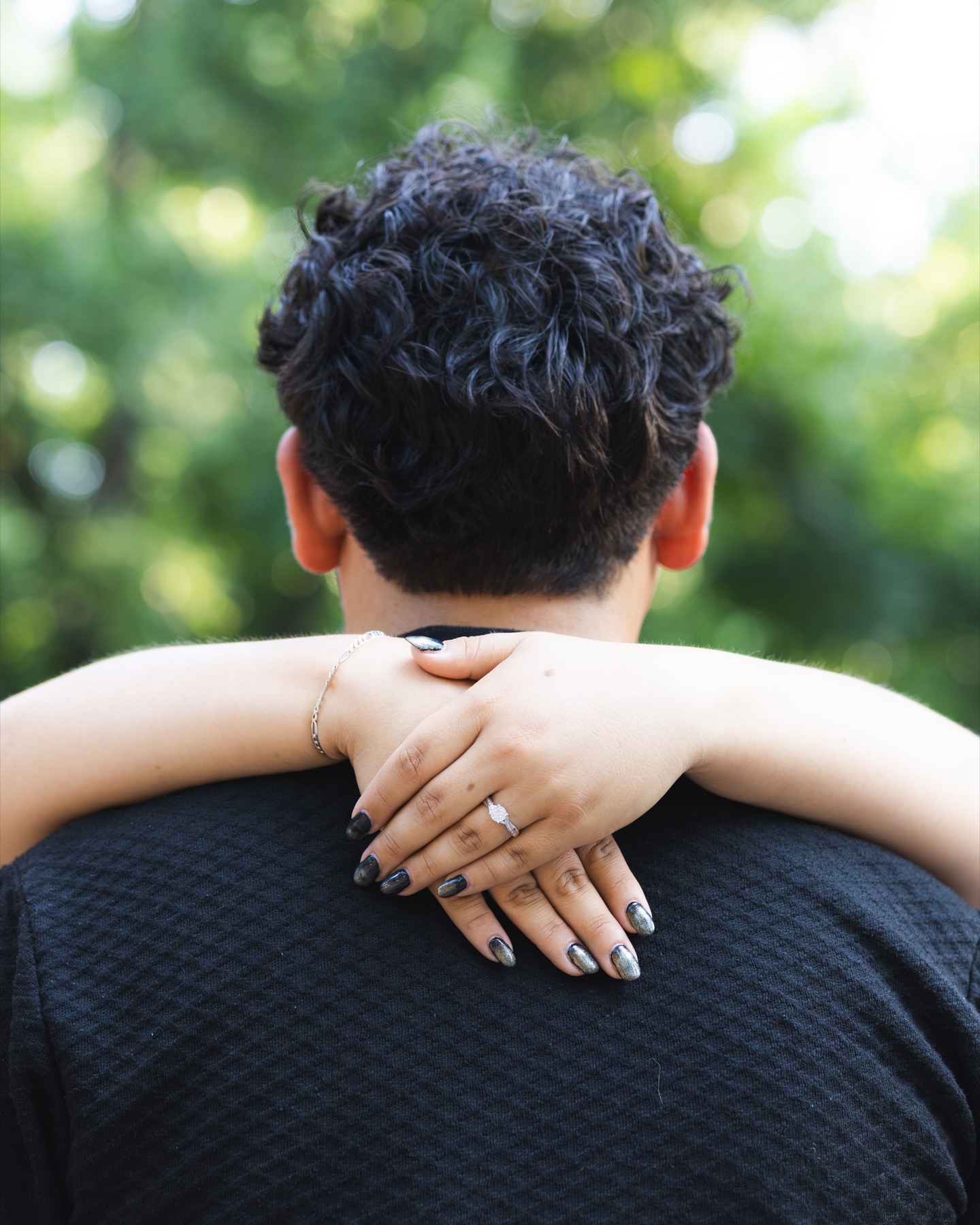 An engagement ring resting on a partner’s shoulder beneath green trees